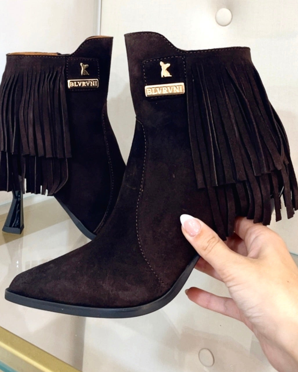 A pair of dark brown suede fringe ankle boots with pointed toes, held by a hand in front of a white tufted display wall.