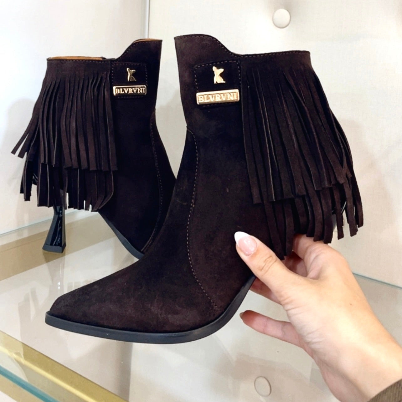 A pair of dark brown suede fringe ankle boots with pointed toes, held by a hand in front of a white tufted display wall.