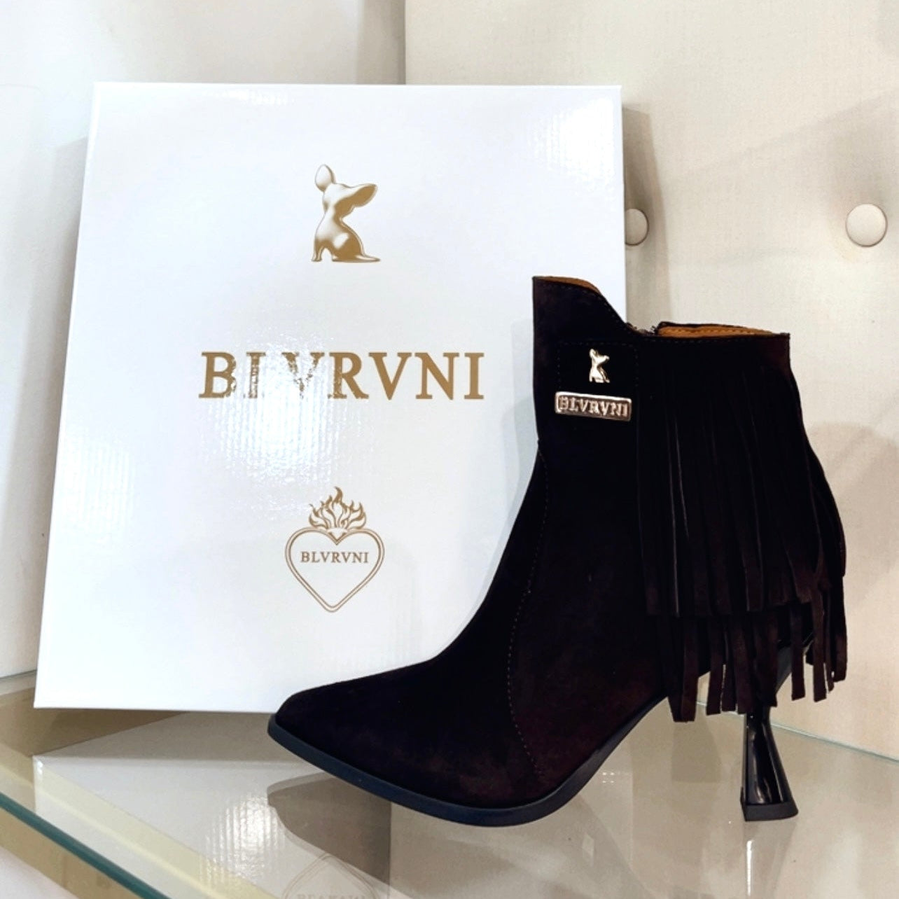 A black suede ankle boot with fringe detailing and a high heel, displayed next to its white BIVRVNI brand box on a glass shelf.