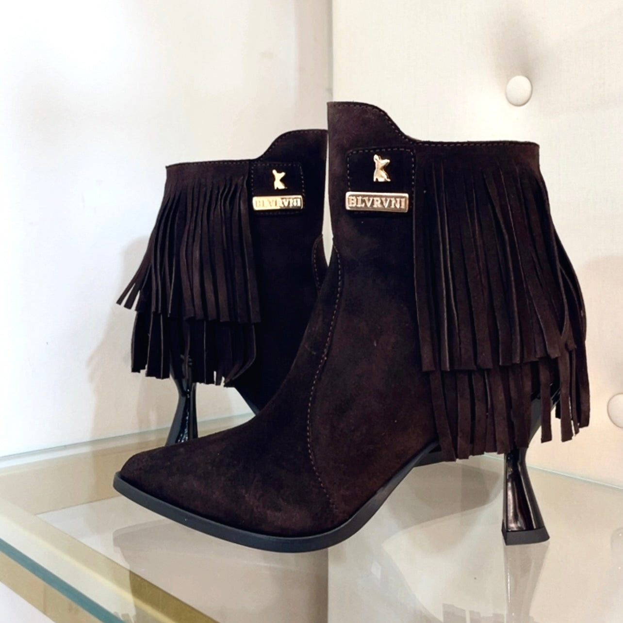 A pair of dark brown suede ankle boots with long fringe detailing and a pointed toe, displayed on a glass shelf against a white tufted wall.