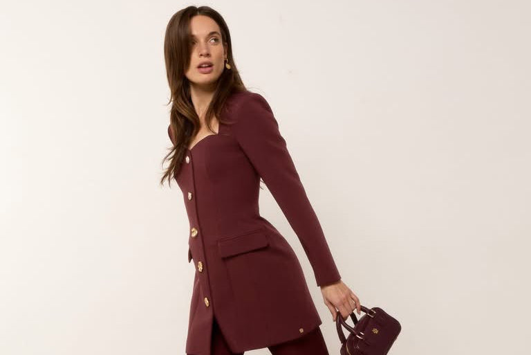 A woman models a matching burgundy two-piece set consisting of a long-sleeve blazer-style mini dress with gold buttons and matching tights, paired with dark pointed-toe heels and a small structured handbag, set against a plain off-white wall and beige carpet.