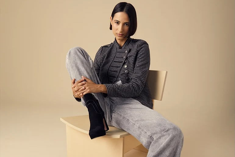 A model sits on a beige geometric stool against a solid beige studio background, wearing a black and white patterned blazer, light gray wide-leg jeans, and black ankle boots.