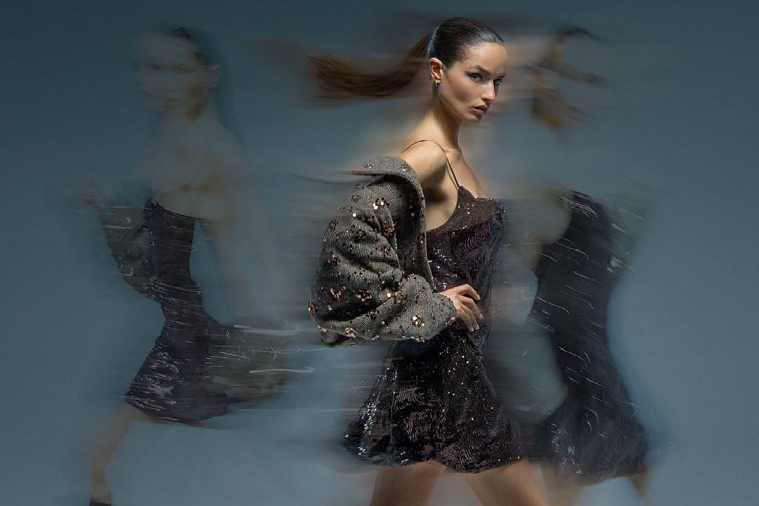 A fashion photograph of a model walking in a studio with motion blur, wearing a dark sequined mini dress, a sparkly cropped jacket, and tall dark brown heeled boots.